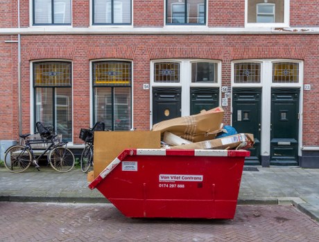 A large red waste skip with a white and red warning label positioned on a paved sidewalk in front of a row of brick terraced houses in Esher, Surrey. Inside the skip, various discarded items are visible, including flattened cardboard boxes, brown paper wrapping, and some plastic packaging, indicating recent rubbish removal activity. To the left of the skip, two bicycles are parked against the brick wall of the homes, with one bicycle's front wheel slightly turned and a piggyback rack attached to its rear. The houses in the background feature tall, narrow sash windows with decorative yellow-tinted glass and brick facades, contributing to a typical residential street scene in Surrey. The scene is well-lit, suggesting it was taken during daylight hours, and the overall setting reflects an urban environment suitable for domestic waste collection and clearance services, highlighting the role of House Clearance Esher in managing local rubbish disposal needs.