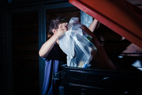 A person is seen loading clear plastic rubbish bags filled with various household waste into a large, black wheelie bin outdoors. The individual, whose face is partially obscured, is wearing a dark casual shirt and is standing on a paved surface adjacent to a wooden fence or shed in a dimly lit environment. The plastic bags contain miscellaneous items, likely including paper, plastic, and other domestic waste materials, indicating a household rubbish removal process. The scene suggests a residential setting, possibly on a driveway or garden area, with natural and artificial light creating shadows along the scene. The image visually aligns with rubbish clearance activities conducted by companies such as House Clearance Esher, servicing areas around Esher and the surrounding postcode, supporting efficient waste collection and disposal services within the local community.