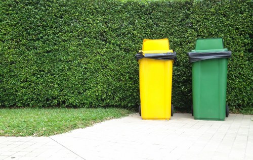 Two wheeled rubbish bins, one yellow and one green, are positioned on a paved area beside a neatly trimmed hedge. The yellow bin is slightly taller and has a black lid with what appears to be a black handle or bag on top. The green bin has a similar black lid, also with a bag or object resting on it. The paved surface consists of rectangular concrete or stone slabs, and the hedge behind the bins is dense and well-maintained, extending across the background. The scene suggests an outdoor setting, likely part of a residential property in Esher, with the bins prepared for rubbish collection. The arrangement of the bins indicates routine waste disposal, aligned with local rubbish removal services provided by House Clearance Esher.