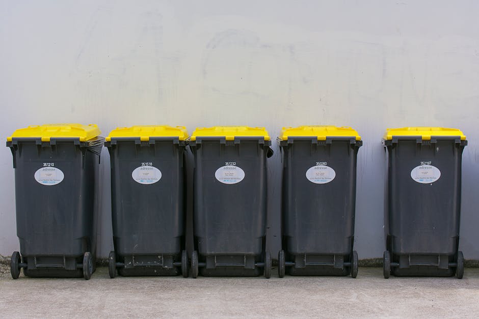 A collection of discarded waste lies on a gravel surface in an outdoor area, including a worn, beige car seat with a fabric covering, black and clear plastic rubbish bags filled with waste, and bright